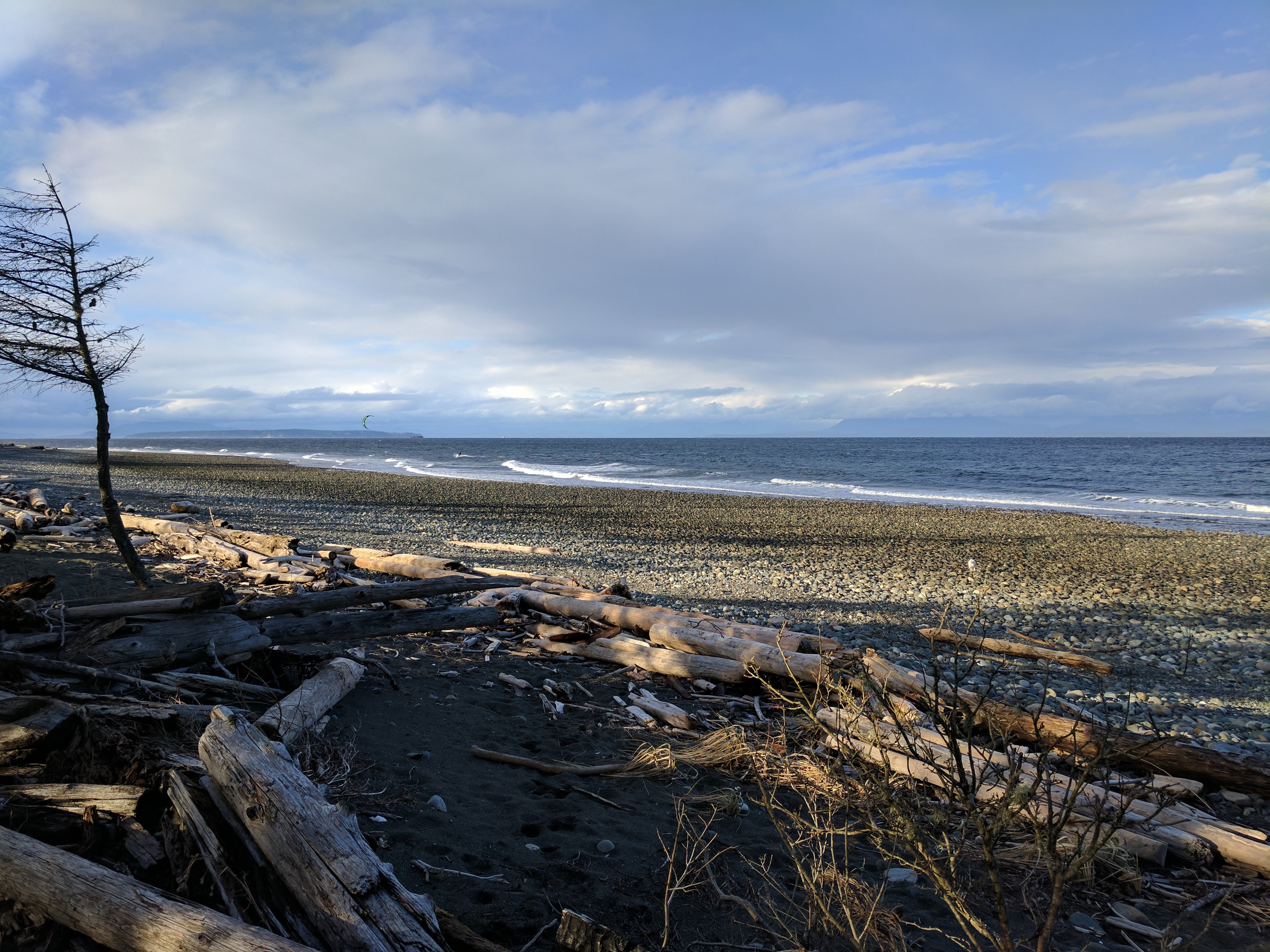 A photograph of a rocky beach on a windy day. A parasailor rides in the distance.