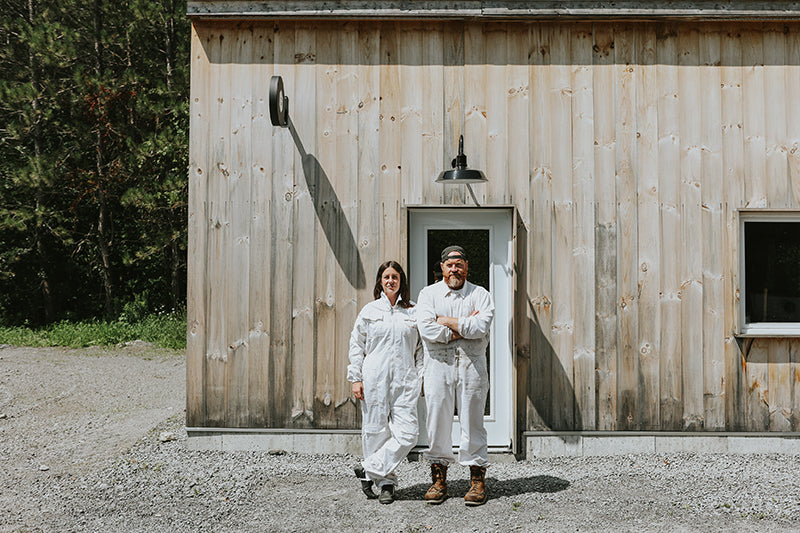 Two people in white coveralls standing in front of a wooden building with a forest background.