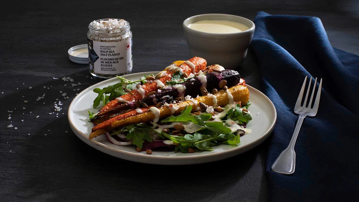 Plated salad with roasted vegetables, a bowl of dressing, and a jar of seasoning on a dark surface.