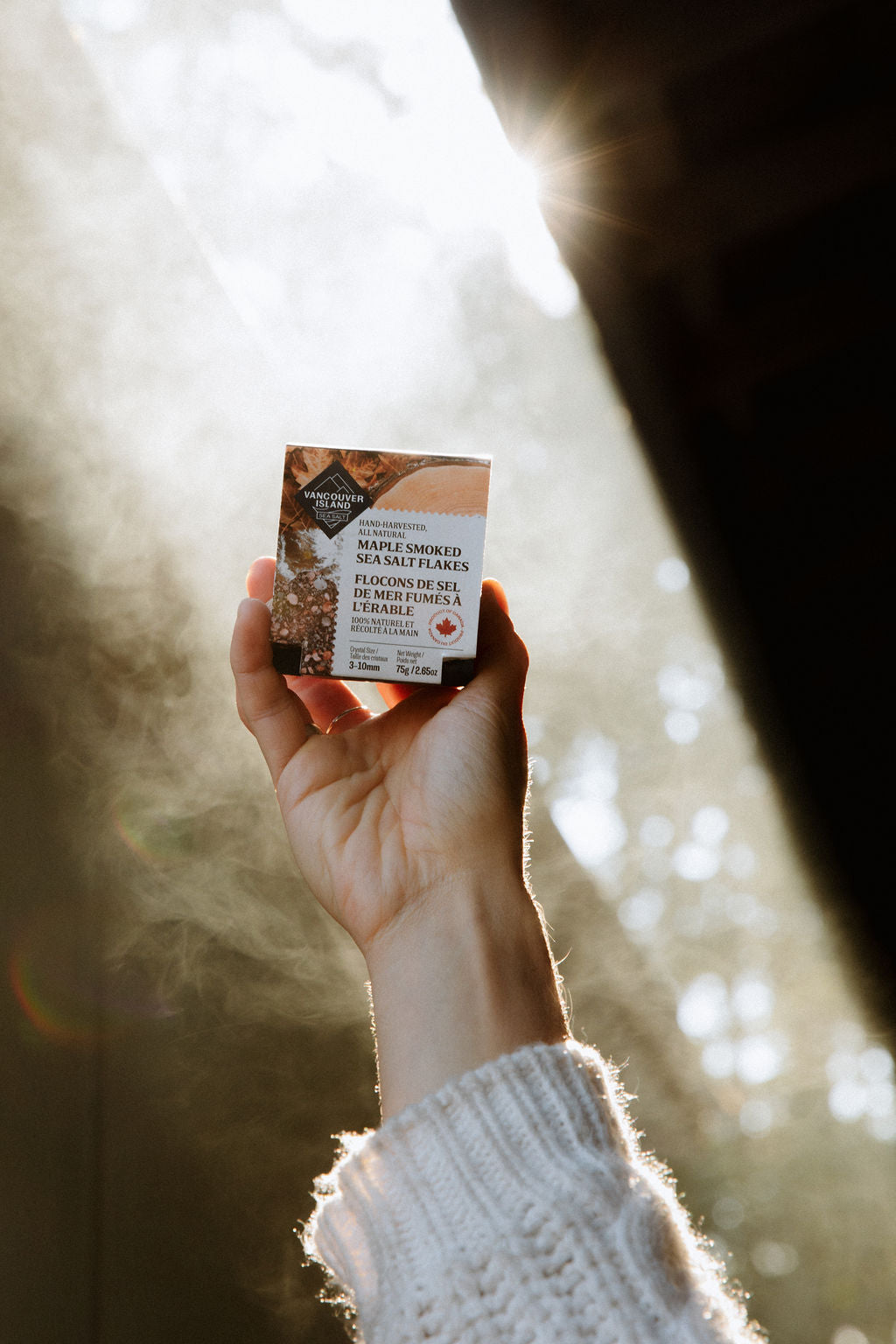 A hand holding Maple Smoked Sea Salt Flakes up to the sun while smoke swirls around