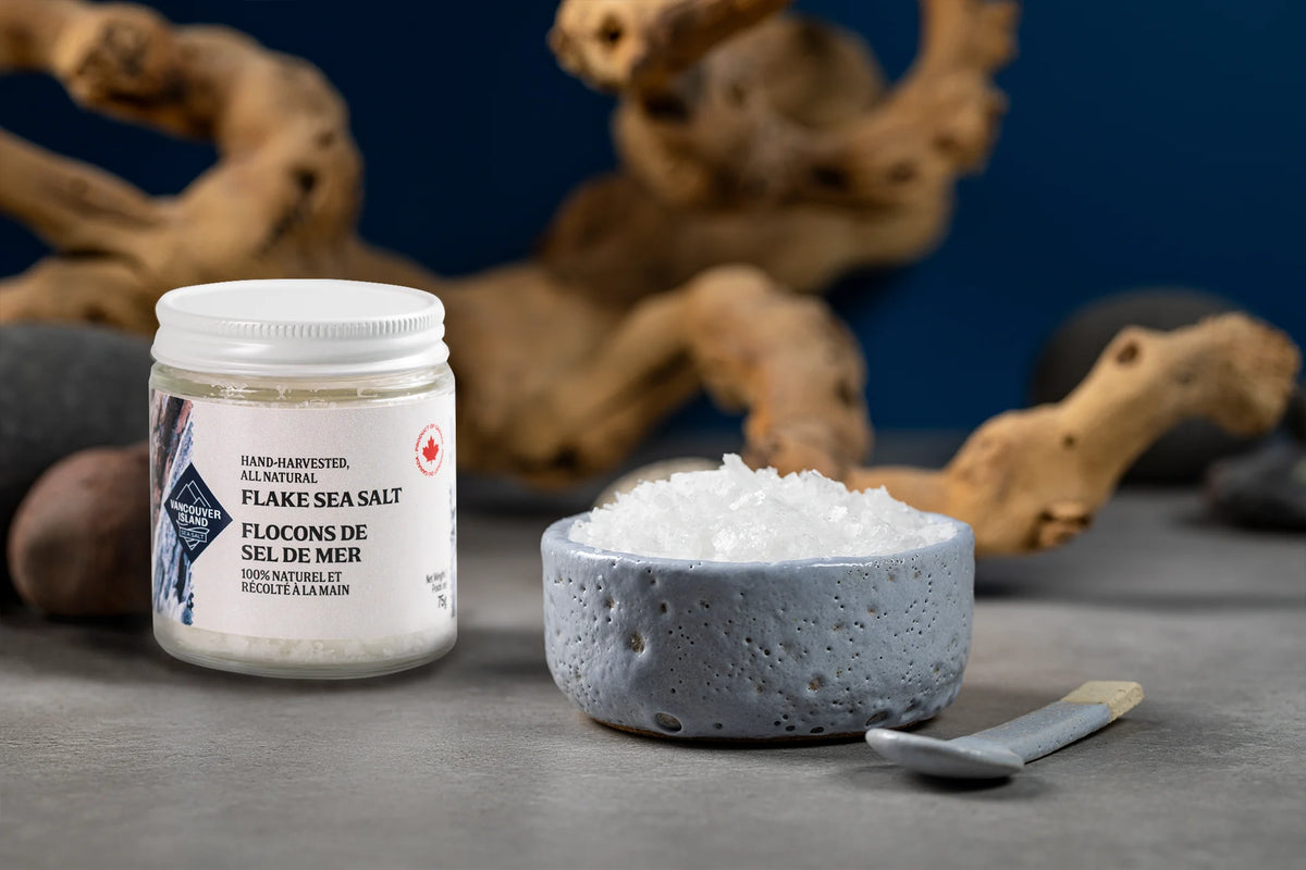 Jar of Vancouver Island Sea Salt flake sea salt beside a ceramic bowl of salt flakes and a small spoon, with driftwood behind