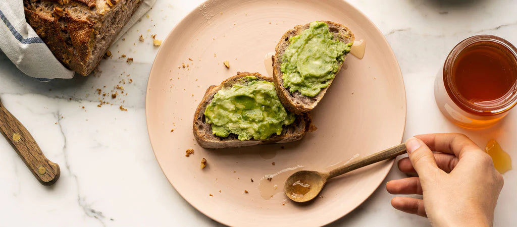 Avocado toast on a plate with a hand reaching for a jar of honey.