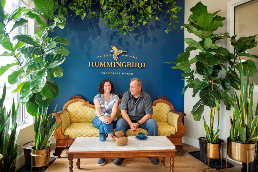Two people sitting on a couch in a room with plants and a 'Hummingbird Chocolate Maker' sign.