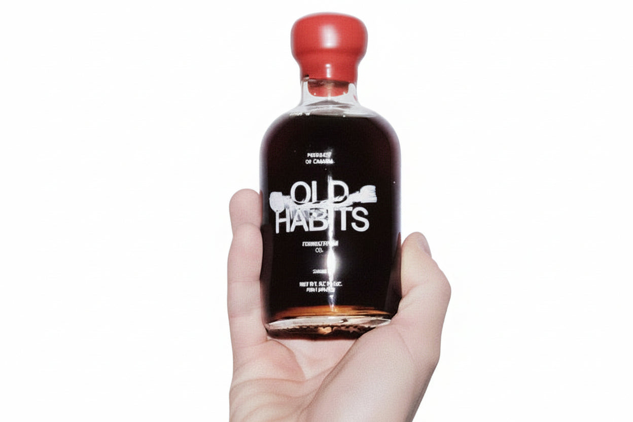 Bottle of 'Old Habits' with a red cap held in a hand against a white background
